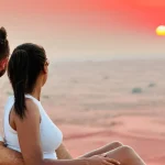 Couple sits together on a sand dune, watching the sunset over a vast desert landscape.