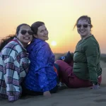 Three people sit together on a sand dune at sunset in the desert, with the sun low on the horizon.