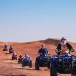 A group of people riding blue quad bikes across orange sand dunes under a clear blue sky.