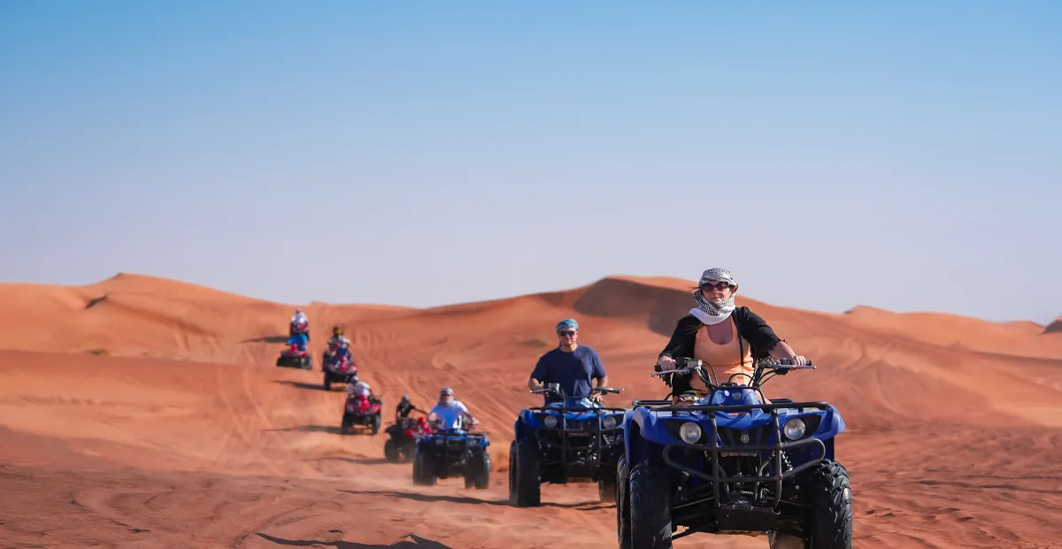 A group of people riding blue quad bikes across orange sand dunes under a clear blue sky.