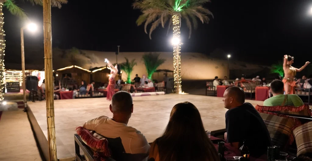 Guests sit on cushioned benches at an outdoor desert camp at night, watching a live belly dance performance on a lit stage surrounded by palm trees wrapped in fairy lights. The sandy dunes and traditional tents are visible in the background, creating an authentic Dubai desert camping atmosphere.