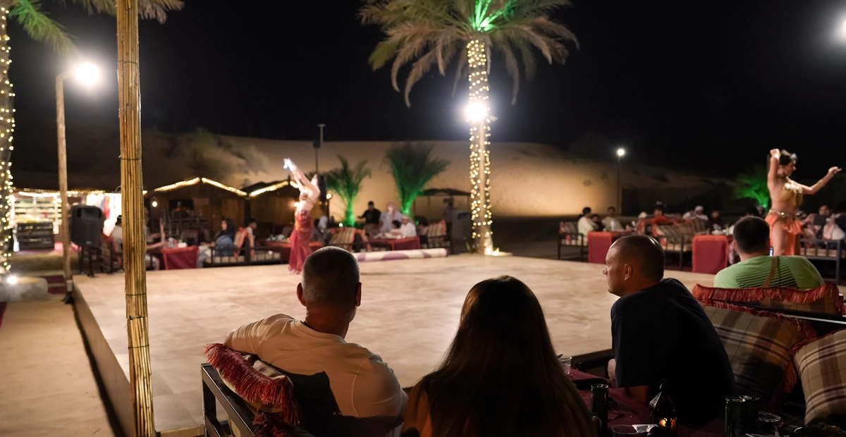 Guests sit on cushioned benches at an outdoor desert camp at night, watching a live belly dance performance on a lit stage surrounded by palm trees wrapped in fairy lights. The sandy dunes and traditional tents are visible in the background, creating an authentic Dubai desert camping atmosphere.