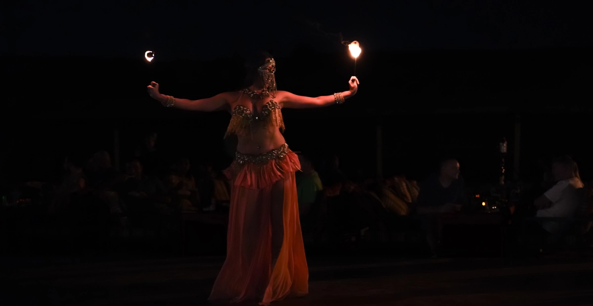 A belly dancer performs at night during a Dubai desert safari. She stands on a dark outdoor stage, holding two lit fire torches outstretched in each hand. She wears a sparkling gold and orange costume with a shimmering skirt and ornate jewelry. The background fades into darkness with silhouettes of seated spectators, capturing a festive and dramatic desert atmosphere.