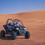 Two people in a blue dune buggy are driving on a large red sand dune under a clear blue sky.