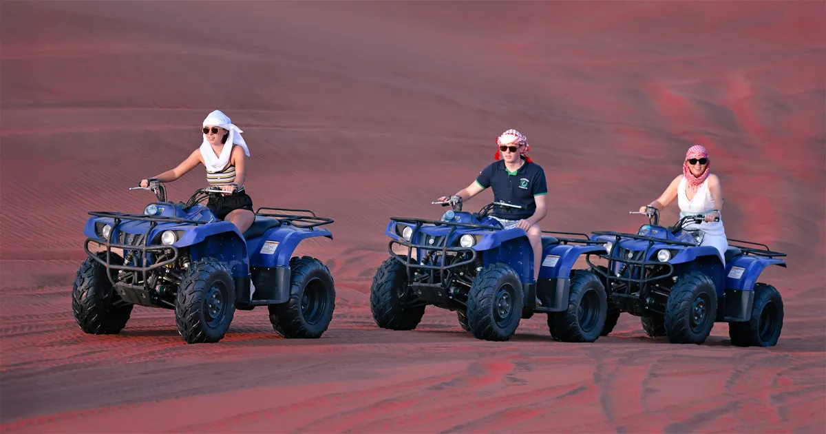 Three people, two women and one man, are riding blue quad bikes across a vast red desert landscape. They are all wearing traditional Middle Eastern head coverings, sunglasses, and casual clothing. The woman on the left wears a white headscarf, a striped top, and black shorts. The man in the middle wears a red and white checkered headscarf, a blue t-shirt, and dark shorts. The woman on the right wears a pink and white patterned headscarf, sunglasses, and a white top.