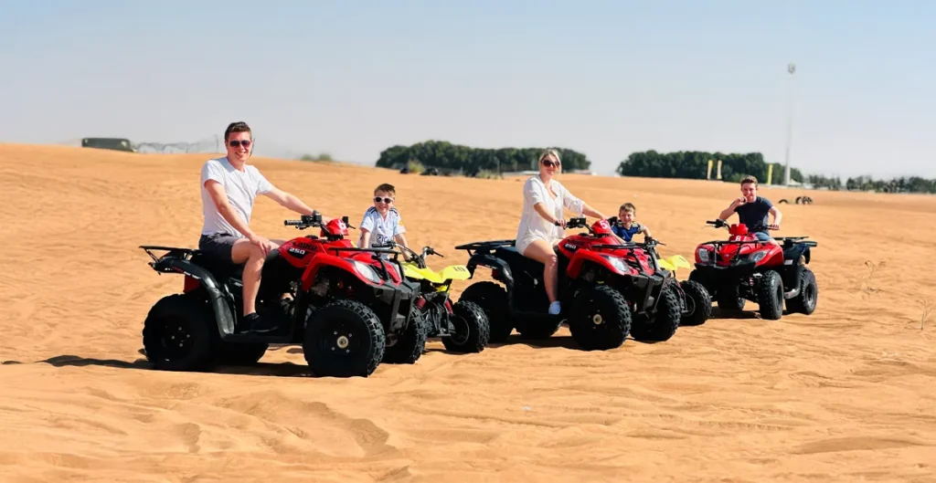 Parents and their toddlers are seated on red and yellow quad bikes in the sand dunes during a desert safari.