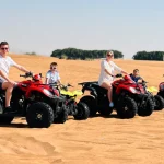 Parents and their toddlers are seated on red and yellow quad bikes in the sand dunes during a desert safari.