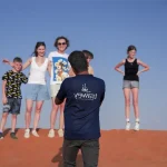 A smiling family of European tourists posing for a photo on a red sand dune, taken by their Trawey guide during a fun desert safari in Dubai.