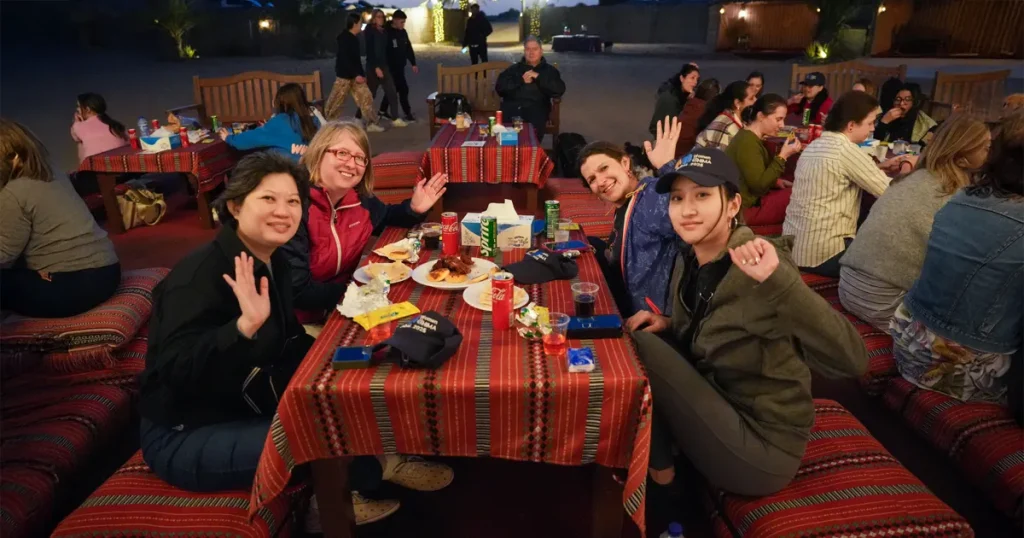 A group of happy tourists smiling and waving while enjoying an evening meal at a traditional desert safari camp in Dubai.