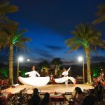 A Tanoura dancer spinning in a vibrant, multi-colored costume on stage at a Trawey desert safari camp in Dubai, viewed by an audience under string lights.
