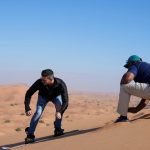 Two men wearing sneakers and outdoor gear walking on sand dunes during a Dubai desert safari.