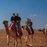 A tour guide in a white thobe leads a caravan of tourists riding camels through the sandy desert dunes under a clear blue sky.