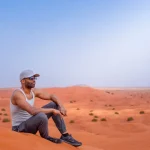 A tourist in a grey tank top sits on a sand dune, enjoying the panoramic view of the Dubai desert landscape during a safari.