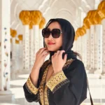 A female tourist wearing a black and gold abaya and headscarf standing among the floral marble columns of Sheikh Zayed Grand Mosque in Abu Dhabi.
