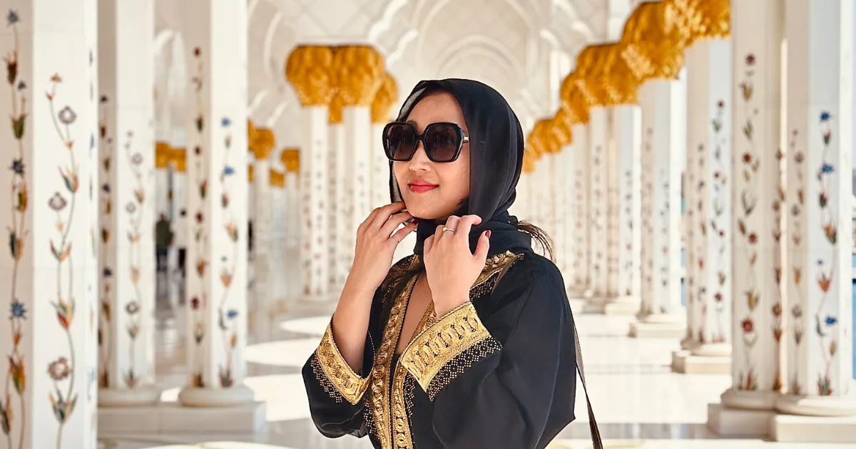 A female tourist wearing a black and gold abaya and headscarf standing among the floral marble columns of Sheikh Zayed Grand Mosque in Abu Dhabi.