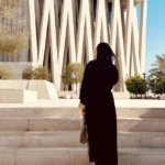 A woman in a traditional black abaya walking up white stone steps toward the striking modern architecture of the Abrahamic Family House in Abu Dhabi.