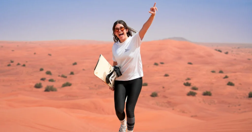 Smiling female tourist cheering while holding a sandboard on a red sand dune during a Dubai desert safari.