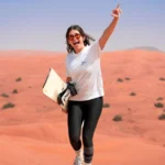 Smiling female tourist cheering while holding a sandboard on a red sand dune during a Dubai desert safari.