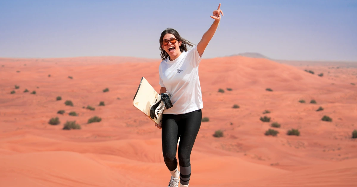 Smiling female tourist cheering while holding a sandboard on a red sand dune during a Dubai desert safari.