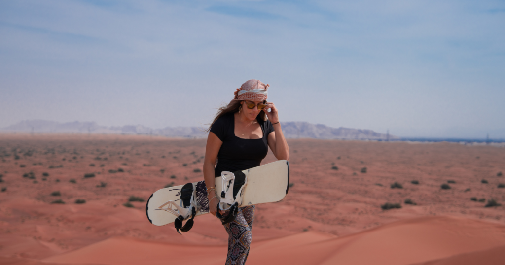 oman wearing protective sunglasses and a headscarf while holding a sandboard on a sunny Dubai desert dune.
