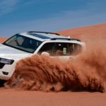 A white Toyota Land Cruiser performing high-speed dune bashing in the Abu Dhabi desert, throwing up a large cloud of red sand.