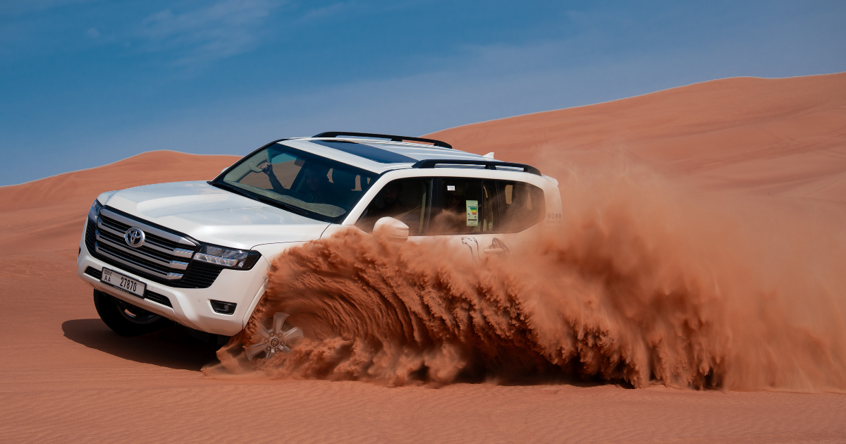 A white Toyota Land Cruiser performing high-speed dune bashing in the Abu Dhabi desert, throwing up a large cloud of red sand.