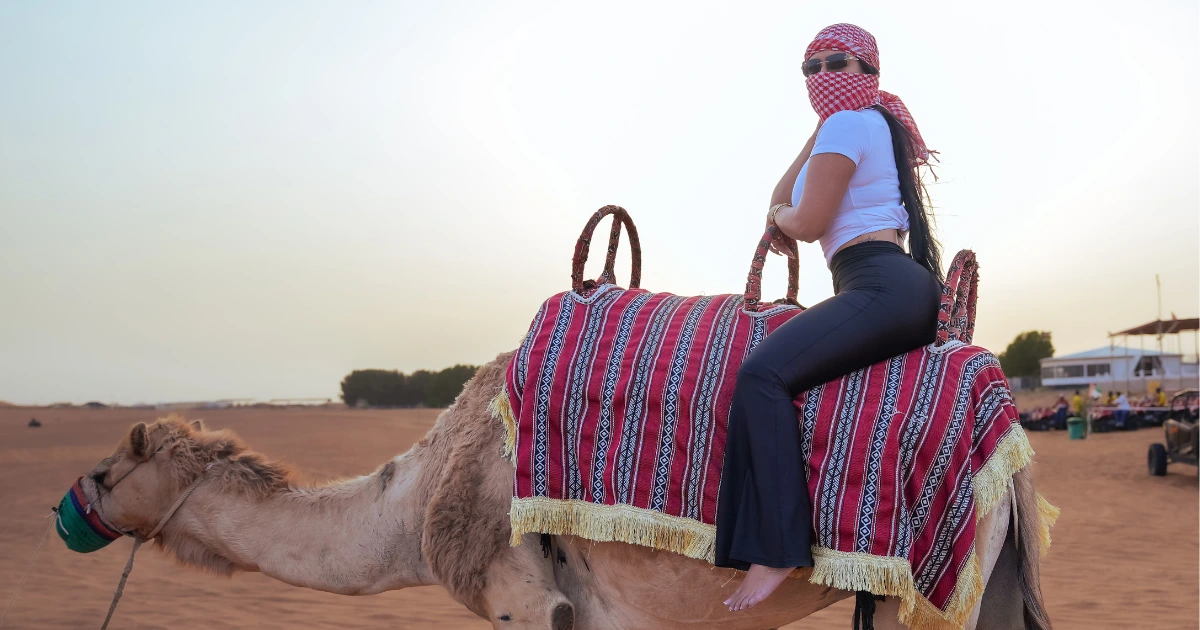 A traveler comfortably enjoying a guided camel ride at sunset, wearing a traditional keffiyeh for an authentic desert experience at a cultural camp.