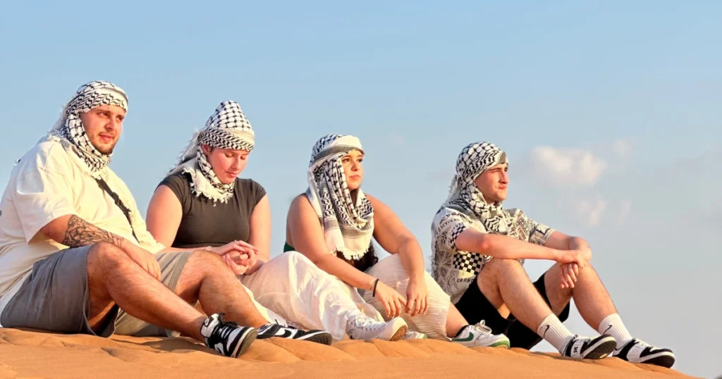 A group of friends wearing traditional keffiyehs sitting peacefully on a sand dune, enjoying a safe and relaxing desert safari experience in Dubai.