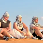 A group of friends wearing traditional keffiyehs sitting peacefully on a sand dune, enjoying a safe and relaxing desert safari experience in Dubai.