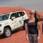 A professional safari guide assisting a tourist with her headscarf next to a white Trawey 4x4 vehicle on a red sand dune.