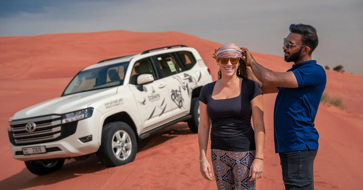 A professional safari guide assisting a tourist with her headscarf next to a white Trawey 4x4 vehicle on a red sand dune.