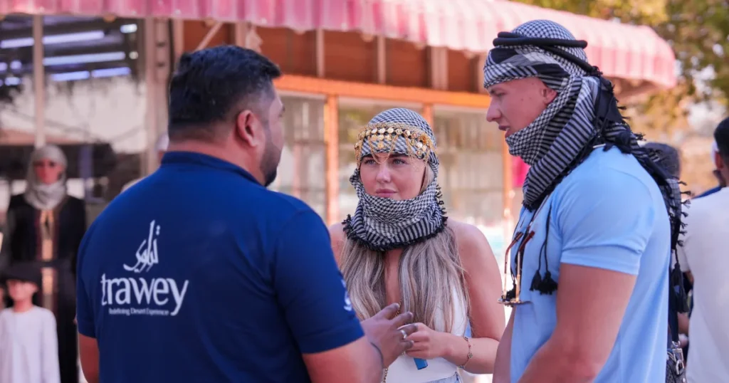 A Trawey safari guide in a blue uniform speaking with two tourists wearing traditional ghutras in a Dubai desert camp.