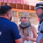 A Trawey safari guide in a blue uniform speaking with two tourists wearing traditional ghutras in a Dubai desert camp.