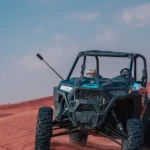 Two people driving a dark grey two-seater dune buggy across red sand dunes.