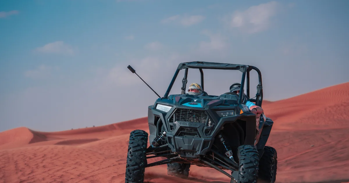 Two people driving a dark grey two-seater dune buggy across red sand dunes.